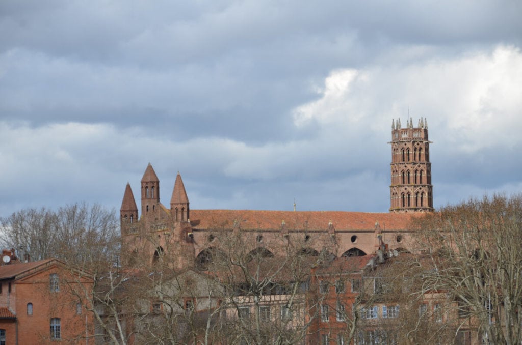 Eine große Backsteinkirche mit mehreren spitzen Türmen und einem hohen Glockenturm erhebt sich über blattlosen Bäumen vor einem wolkenverhangenen Himmel - ein ikonischer Anblick bei einer Stadtbesichtigung in Toulouse, Südfrankreich.