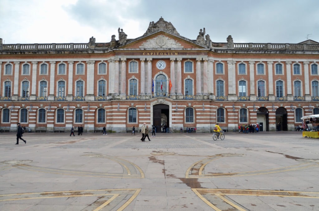 Ein großes, verziertes Gebäude mit Säulen und einer Uhr über dem Haupteingang steht auf einem großen Platz in Toulouse. Menschen spazieren und ein Radfahrer fährt über den offenen Platz unter einem bewölkten Himmel und fängt den Charme der Stadtbesichtigung in Südfrankreich ein.