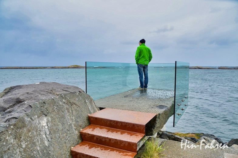 Eine Person in einer grünen Jacke steht auf einer gläsernen Aussichtsplattform über dem Wasser und blickt unter bewölktem Himmel auf den Ozean entlang der Atlantikstraße mit dem Wohnmobil. Nasse, orangefarbene Stufen führen von den Felsen hinauf; ferne Inseln runden das dramatische Bild ab.