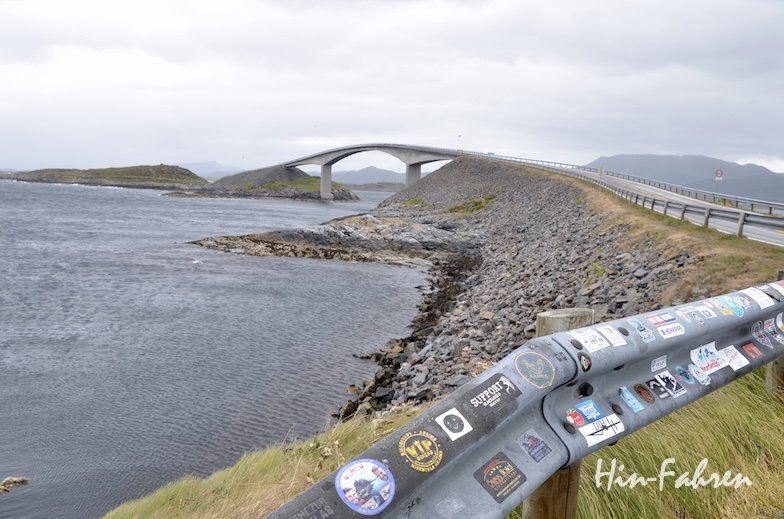 Eine geschwungene Brücke überspannt das Wasser mit seinen felsigen Ufern unter einem bewölkten Himmel. Die Leitplanke im Vordergrund ist mit bunten Aufklebern übersät, während ein Wohnmobil die berühmte Atlantikstraße entlangfährt. In der Ferne sind Hügel und Inseln zu sehen.