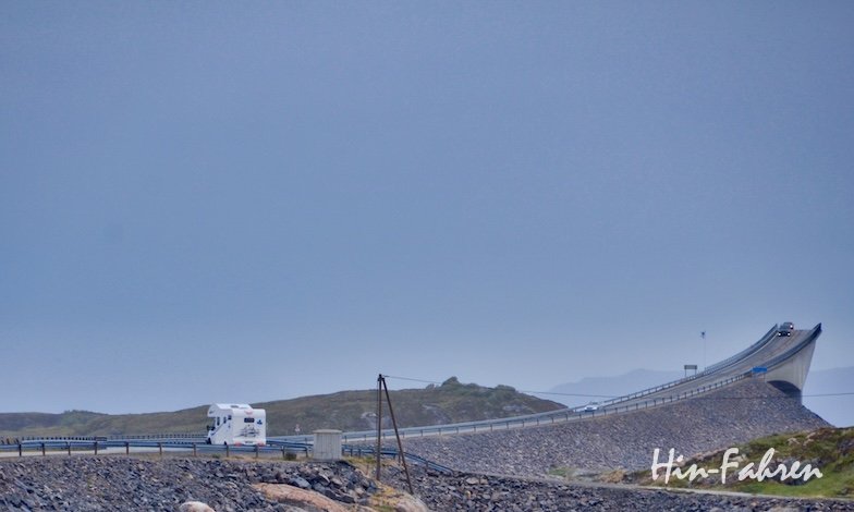 Ein Wohnmobil fährt auf der Atlantikstraße auf eine steile, kurvenreiche Brücke zu und schlängelt sich unter bewölktem Himmel eine felsige Küstenstraße entlang. Die Szene wirkt abgelegen und dramatisch.