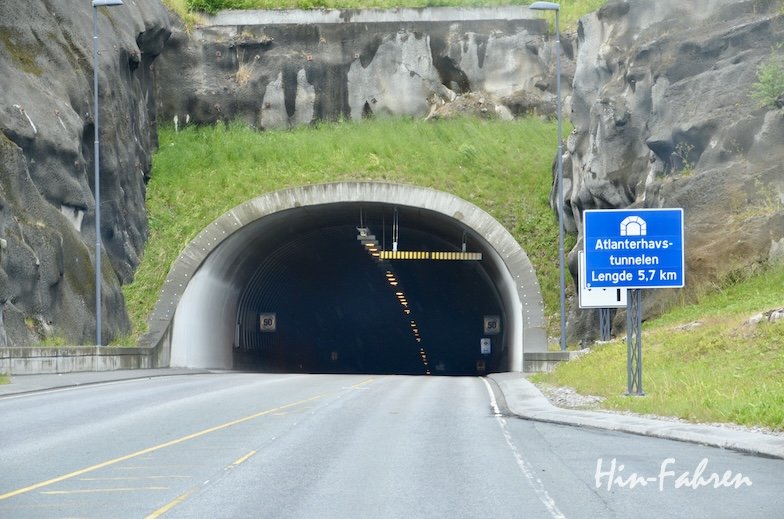 Das Bild zeigt den Eingang zum Atlanterhavstunnelen auf der berühmten „Atlantikstraße mit Wohnmobil“-Route, der sich durch felsiges Gelände schlängelt. Ein blaues Schild weist auf seine Länge von 5,7 km hin; die beleuchtete Tunnelstraße ist leer und am Eingang von Gras und Felsen umgeben.