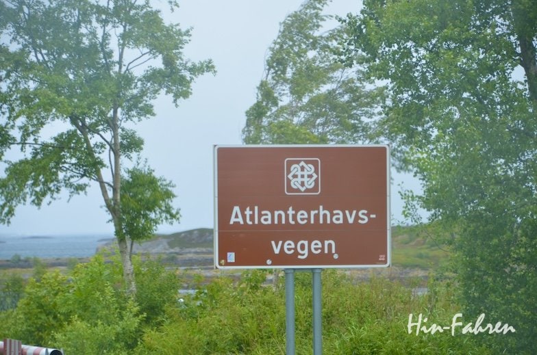 Ein braunes Straßenschild mit weißer Beschriftung trägt die Aufschrift „Atlanterhavsvegen“ und steht inmitten grüner Bäume und Laub, im Hintergrund sind ein bewölkter Himmel und Wasser zu sehen – ideal, um die Atlantikstraße mit dem Wohnmobil zu erkunden.