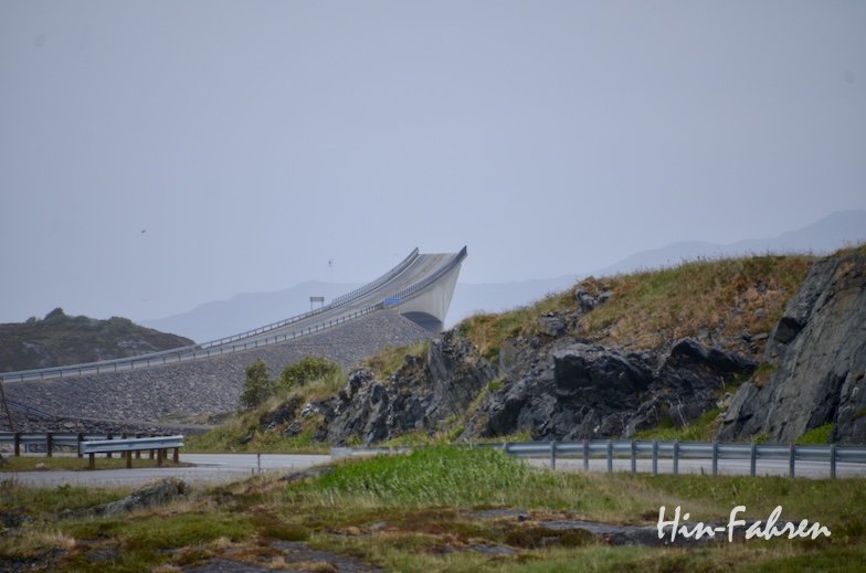 Eine geschwungene Küstenbrücke, die Teil der Route „Atlantikstraße mit Wohnmobil“ ist, erhebt sich über felsigem Gelände, mit Grasflächen und fernen Bergen unter einem bewölkten Himmel. Das Foto trägt unten rechts den Text „Hin-Fahren“.
