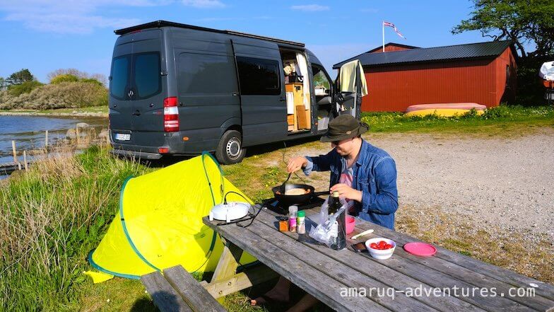 Mann sitzt auf einer Picknickbank und kocht, dahinter ein Van