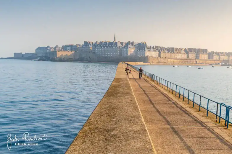 Ein langer steinerner Pier ragt ins Wasser in Richtung einer historischen Stadtmauer mit vielen Gebäuden und einem Kirchturm; an einem klaren, sonnigen Tag spazieren oder radeln ein paar Leute den Pier entlang.