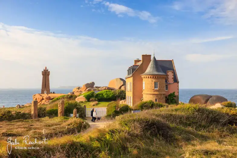 Ein charmantes Steinhaus mit einem Türmchen steht in der Nähe der felsigen Küste, mit einem hohen Leuchtturm und großen rosafarbenen Granitblöcken im Hintergrund unter einem teilweise bewölkten Himmel. Im Vordergrund gehen zwei Personen einen Weg entlang.