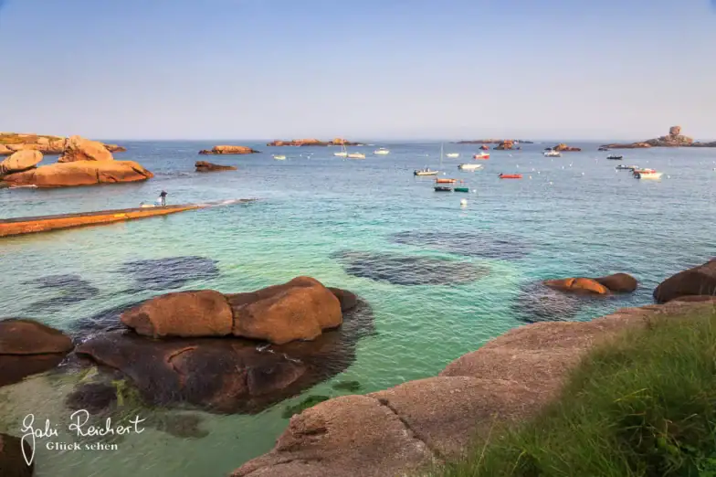 Eine malerische Küstenansicht mit klarem türkisfarbenem Wasser, großen braunen Felsen im Vordergrund und zahlreichen kleinen Booten, die auf dem ruhigen Meer unter einem klaren Himmel schwimmen. Eine Person steht auf einem ins Wasser ragenden Steg.