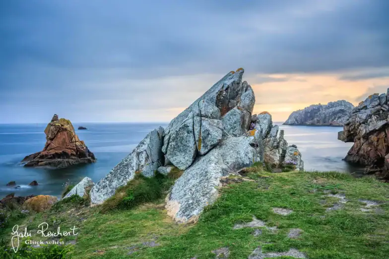 Große, zerklüftete Felsen erheben sich aus dem grünen Gras einer Küstenklippe mit Blick auf ruhiges, blaues Wasser, mit weiteren Felsen in der Ferne unter einem bewölkten Himmel bei Sonnenuntergang.