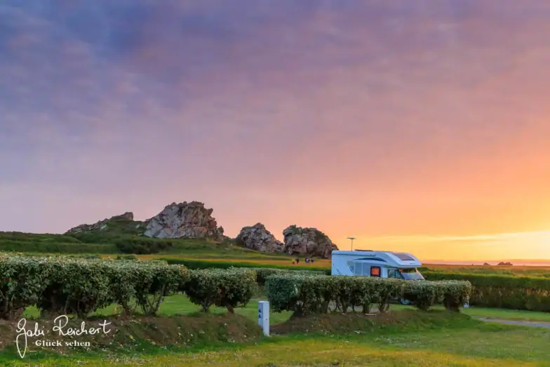 Ein Wohnmobil ist auf einer Wiese in der Nähe von gepflegten Hecken geparkt, mit felsigen Hügeln im Hintergrund und einem leuchtend orangefarbenen und violetten Sonnenuntergangshimmel über uns. Die Szene wirkt friedlich und malerisch.