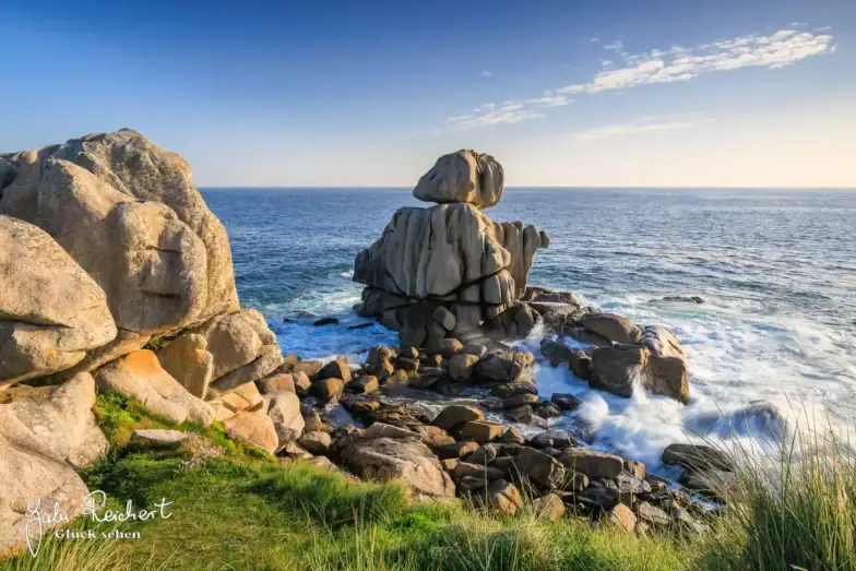 Felsige Küstenlandschaft mit großen Felsbrocken, die in Meeresnähe aufgeschichtet sind, Wellen, die gegen die Felsen schlagen, grünes Gras im Vordergrund und ein klarer blauer Himmel darüber.