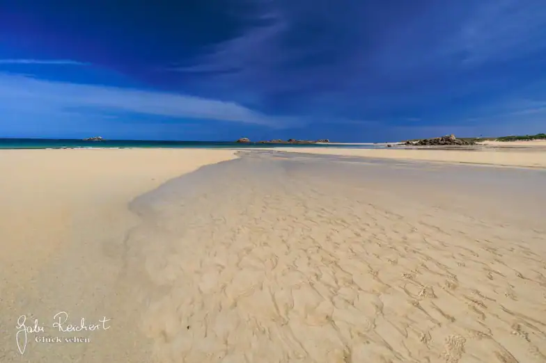 Ein breiter Sandstrand mit sanften Wellen im Sand, seichtes Wasser, in dem sich der blaue Himmel spiegelt, und ein tiefblauer Ozean, der sich unter einem leuchtenden Himmel mit Wolkenfetzen bis zum Horizont erstreckt.