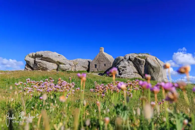 Ein kleines Steinhaus liegt zwischen großen Felsbrocken auf einem grasbewachsenen Hügel, mit Wildblumen im Vordergrund und einem strahlend blauen Himmel im Hintergrund.