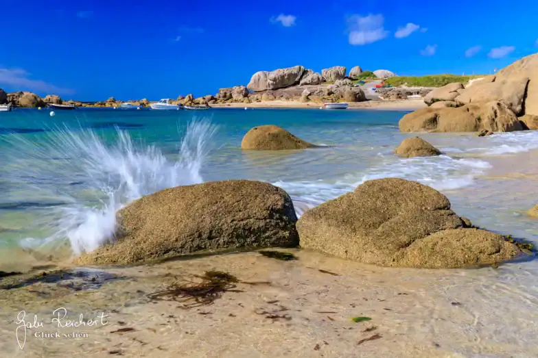 An einem sonnigen Strand mit blauem Himmel, Felsen, Booten in der Ferne und grünem Gras im Hintergrund schlagen die Wellen gegen große Felsen im flachen, klaren Wasser.