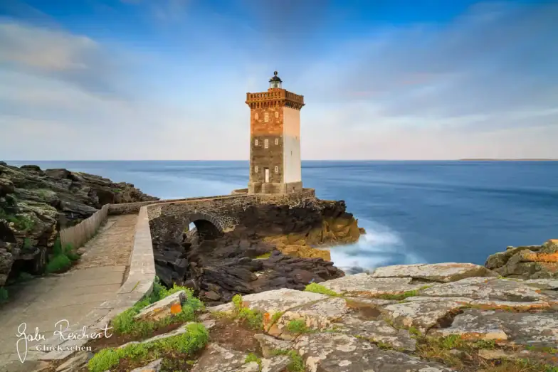 Ein steinerner Leuchtturm steht auf felsigen Klippen am Meer, die durch einen schmalen Steinsteg verbunden sind, unter einem teilweise bewölkten blauen Himmel bei Sonnenuntergang. Die Wellen schlagen sanft gegen die Felsen.
