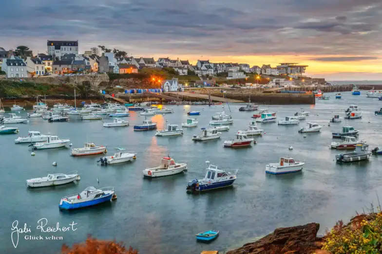 Ein malerischer Hafen bei Sonnenuntergang mit zahlreichen kleinen Booten, die im ruhigen Wasser vor Anker liegen, bunten Häusern und Gebäuden am Ufer und einem dramatisch bewölkten Himmel über der Stadt.