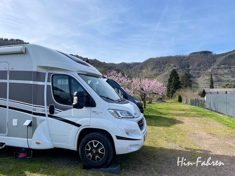 Ein weißes Wohnmobil, das auf einer Wiese neben einem anderen Fahrzeug geparkt ist, mit blühenden rosa Bäumen und grünen Hügeln im Hintergrund unter einem blauen Himmel. Die Worte Hin-Fahren erscheinen in der unteren rechten Ecke.