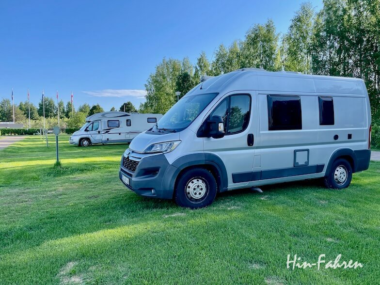 Ein silbernes Wohnmobil steht auf einem grasbewachsenen Campingplatz unter blauem Himmel, mit einem anderen Wohnmobil im Hintergrund und Bäumen, die das Gebiet umgeben. Der Schriftzug Hin-Fahren erscheint in der unteren rechten Ecke.