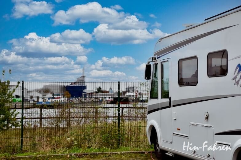 Ein weißes Wohnmobil ist an einem Metallzaun geparkt, mit Blick auf einen Hafen mit Booten und Gebäuden unter einem teilweise bewölkten Himmel.