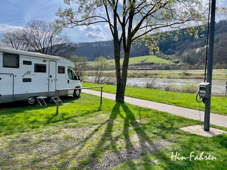 Ein Wohnmobil ist auf einer Wiese in der Nähe eines Flusses geparkt, mit einem Picknicktisch und einem Baum, der lange Schatten wirft. Im Hintergrund sind Hügel und Bäume unter einem teilweise bewölkten Himmel zu sehen.