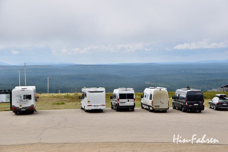 Mehrere Wohnmobile und Wohnwagen sind in einer Reihe am Rande eines Grundstücks geparkt, mit Blick auf eine weite, bewaldete Landschaft mit fernen Hügeln unter einem bewölkten Himmel. In der rechten unteren Ecke ist das Hin-Fahren-Logo zu sehen.