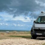 Ein silbernes Wohnmobil steht auf einer Schotterfläche in der Nähe von grasbewachsenen Dünen mit Blick auf ein wolkenverhangenes, windiges Meer unter einem dramatischen Himmel.