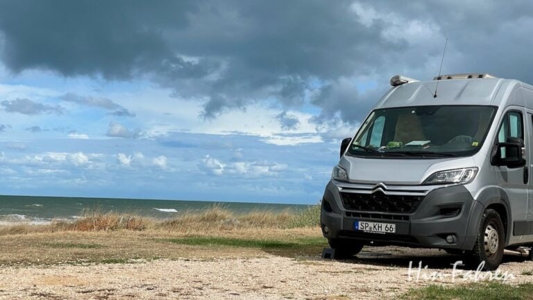 Ein silbernes Wohnmobil steht auf einer Schotterfläche in der Nähe von grasbewachsenen Dünen mit Blick auf ein wolkenverhangenes, windiges Meer unter einem dramatischen Himmel.