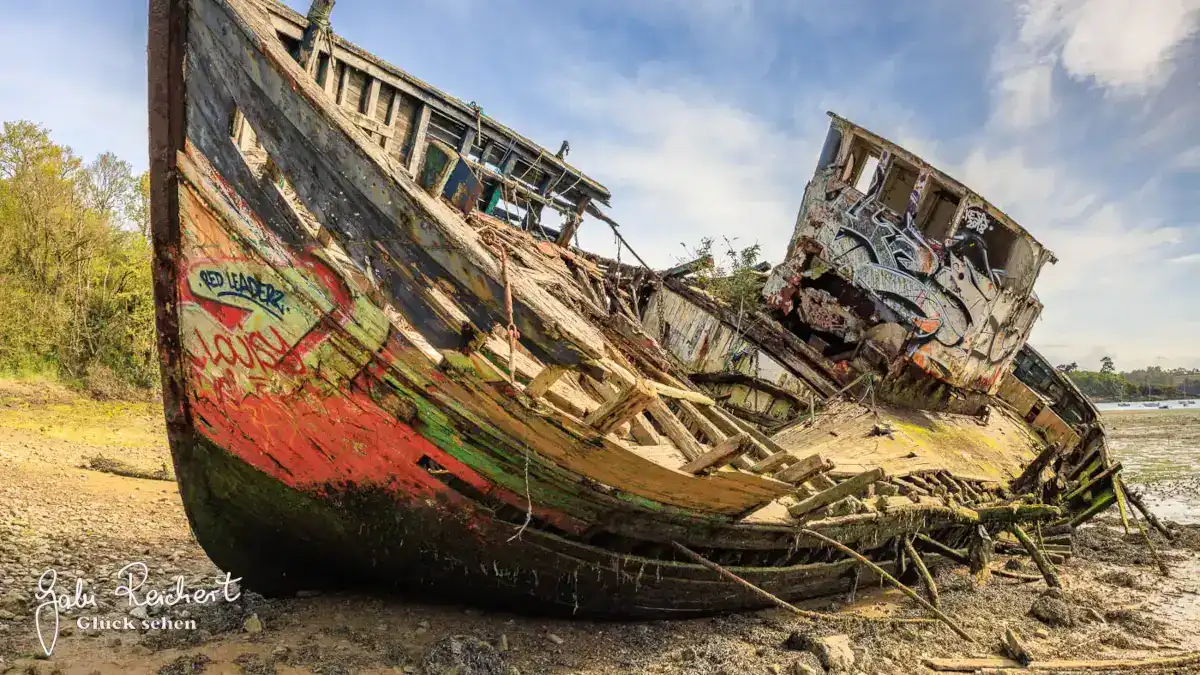 Ein verfallendes, mit Graffiti beschmiertes hölzernes Schiffswrack liegt an einem sandigen, schlammigen Ufer unter blauem Himmel. Die Farbe des Schiffsrumpfs blättert ab und legt grünes, rotes und natürliches Holz frei, Teile stürzen ein und in der Nähe wachsen Pflanzen.