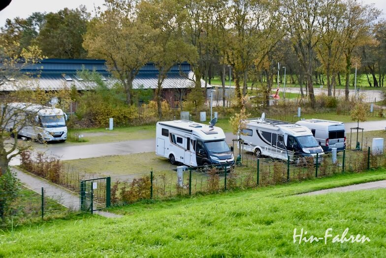 Vier Wohnmobile stehen auf gepflasterten Plätzen in einem kleinen, eingezäunten Campingplatz, umgeben von Gras und Bäumen mit Herbstlaub. Die Szene wirkt ruhig, im Hintergrund sind eine Straße und ein Gebäude zu sehen.