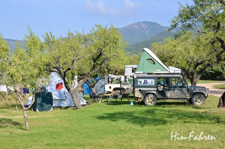 Ein grüner Pop-up-Camper und Zelte sind unter Bäumen auf einem grasbewachsenen Campingplatz aufgebaut, mit Bergen im Hintergrund und verstreutem Campingzubehör.
