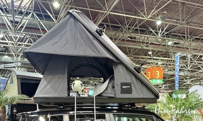 Ein schwarzes Dachzelt mit dreieckigem Muster ist auf einem Fahrzeug in einer großen überdachten Ausstellungshalle montiert, wobei im Hintergrund Metallträger und Schilder zu sehen sind.