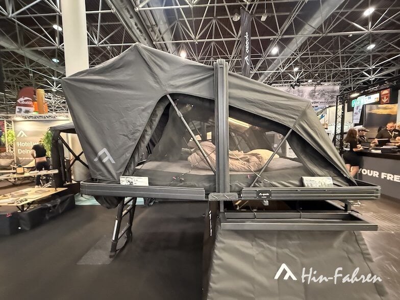 Ein großes überdachtes Zelt wird in einem Ausstellungsraum ausgestellt. Es ist auf einem Metallrahmen mit schwarzem Stoff, Fenstern mit Reißverschluss und Netzpaneelen aufgebaut. Im Hintergrund sind Menschen und verschiedene Stände zu sehen.