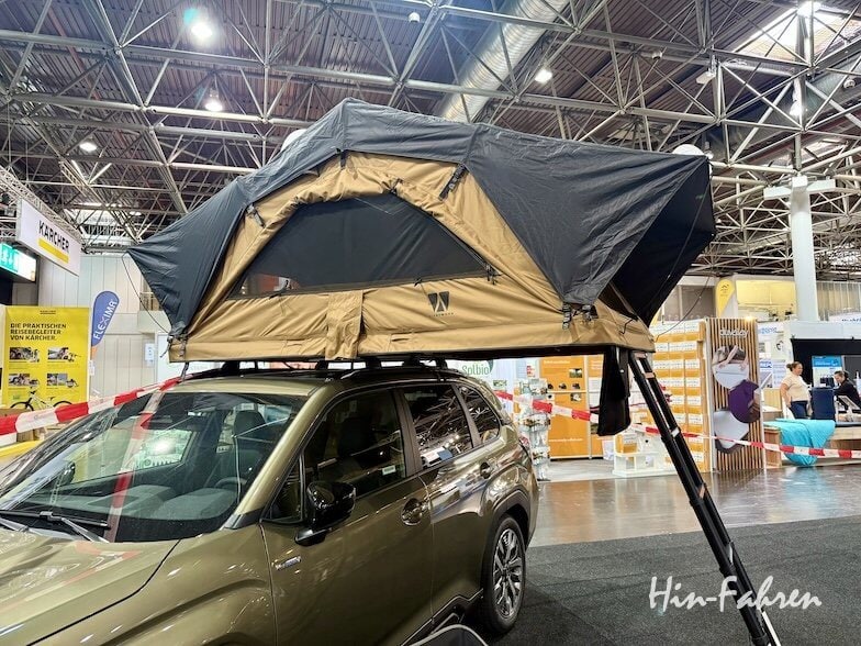 Ein brauner Geländewagen mit einem großen, auf dem Dach montierten Zelt ist in einem Ausstellungsraum ausgestellt. Das Zelt ist mit Fenstern erweitert, und eine Leiter reicht vom Zelt bis zum Boden.