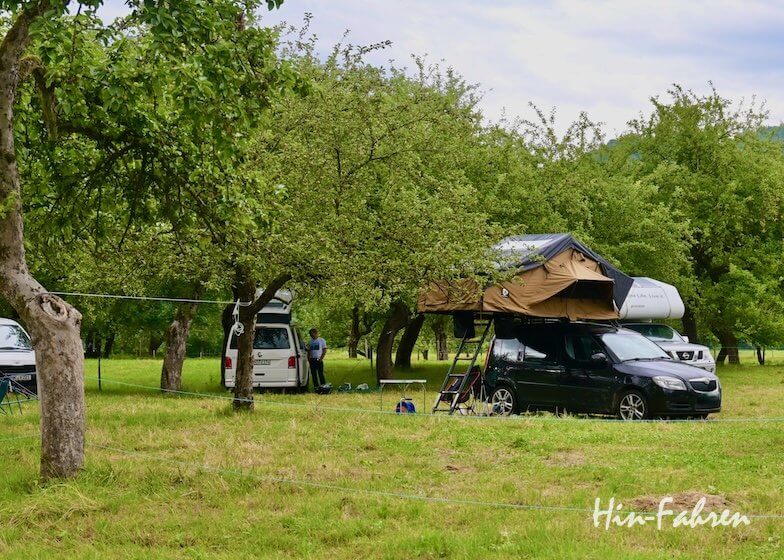 Zwei Wohnmobile sind auf einer Wiese zwischen Bäumen geparkt, eines davon mit einem Zelt auf dem Dach. Campingstühle und -ausrüstung sind zu sehen, und zwei Personen stehen in der Nähe des einen Wagens. Die Szene ist ruhig und im Freien angesiedelt. Der Hin-Fahren-Text steht in der Ecke.