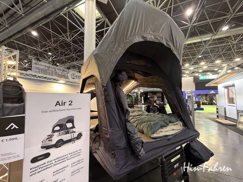 Ein auf einem Fahrzeug aufgebautes Dachzelt wird auf einer Indoor-Ausstellung gezeigt. Im Inneren des Zeltes ist ein Schlafsack zu sehen. Auf einem Schild neben dem Aufbau sind Produktdetails und Preise angegeben. Im Hintergrund sind andere Stände zu sehen.