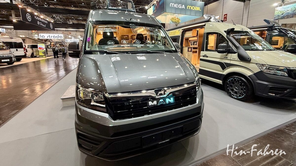 Ein graues MAN-Wohnmobil wird auf einer Indoor-Ausstellung ausgestellt, wobei die Lichter auf seiner Motorhaube reflektiert werden. Im Hintergrund sind weitere Wohnmobile und Messestände zu sehen. Das Bild ist mit dem Wasserzeichen Hin-Fahren versehen.