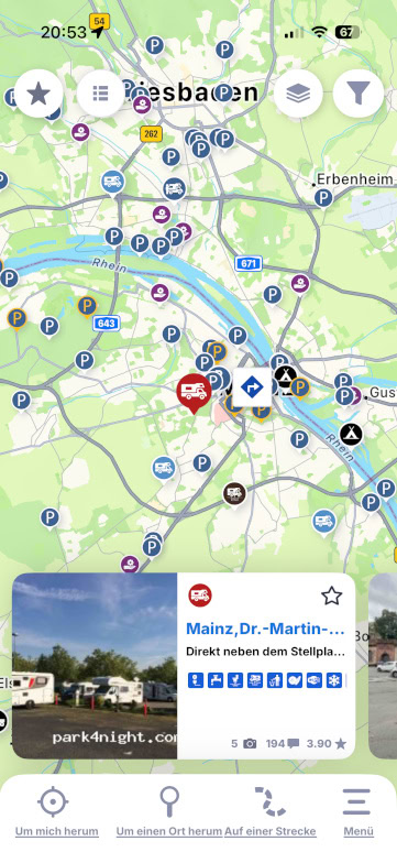 Eine Karten-App für Smartphones zeigt Wiesbaden und Mainz mit Symbolen für Parkplätze, Campingplätze und Dienstleistungen. Ein ausgewählter Standort in der Nähe des Rheins wird mit einem Foto und Details zu einem Parkplatz in Mainz angezeigt.