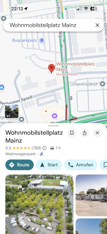 Ein Smartphone-Screenshot zeigt eine Google-Maps-Suche nach dem Wohnmobilstellplatz Mainz mit einer Karte, einer Standortnadel, einer Bewertung und Bildern eines Wohnmobilstellplatzes mit mehreren geparkten Fahrzeugen und umliegenden Grünflächen.