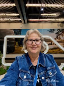 Eine Frau mit kurzen grauen Haaren und Brille lächelt in die Kamera. Sie trägt eine blaue Jeansjacke und ein Schlüsselband. Hinter ihr ist eine malerische Kulisse mit Bäumen, Wasser und einem Wohnmobil zu sehen.