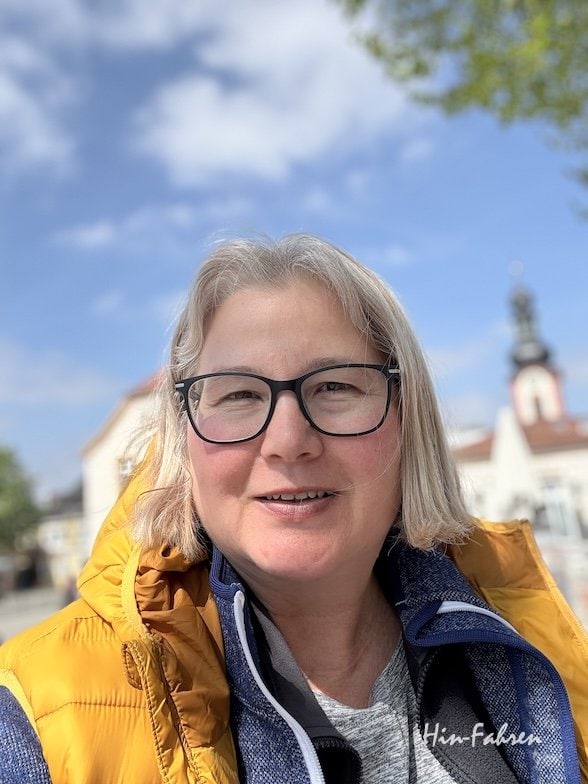 Eine Frau mit kurzen grauen Haaren und Brille lächelt in die Kamera. Sie trägt eine blaue Jeansjacke und ein Schlüsselband. Hinter ihr ist eine malerische Kulisse mit Bäumen, Wasser und einem Wohnmobil zu sehen.