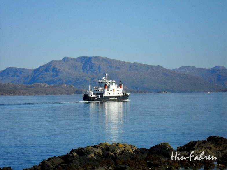 Eine schwarz-weiße Fähre fährt auf ruhigem blauem Wasser mit felsigen Hügeln und Bergen im Hintergrund unter einem klaren Himmel. Im Vordergrund ist eine felsige Küste zu sehen.