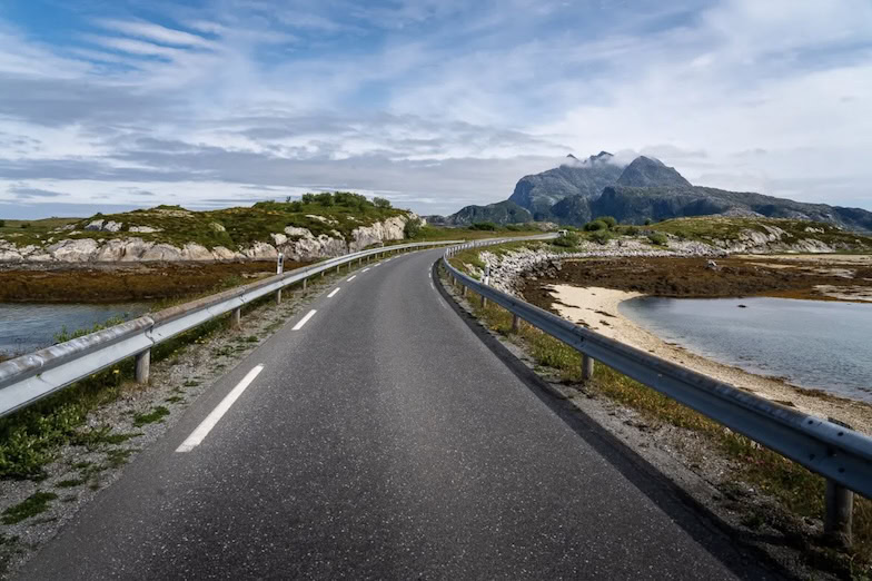 Eine kurvenreiche Straße mit Metallgeländern führt durch felsiges Küstengelände an einem ruhigen Gewässer entlang. Im Hintergrund erheben sich grüne Hügel und ein Berg unter einem teilweise bewölkten Himmel.
