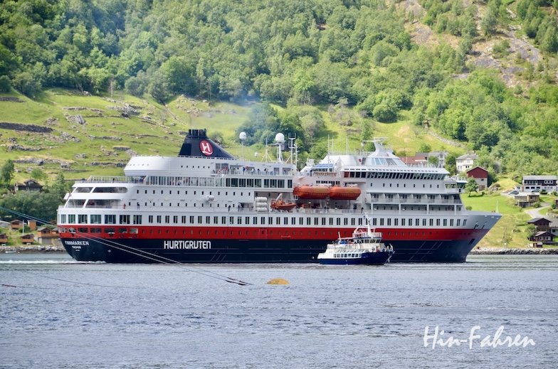 Ein großes Hurtigruten-Kreuzfahrtschiff fährt in der Nähe eines norwegischen Dorfes, daneben liegt ein kleineres Schiff. Im Hintergrund sind grüne Hügel und verstreute Häuser unter einem teilweise bewölkten Himmel zu sehen.