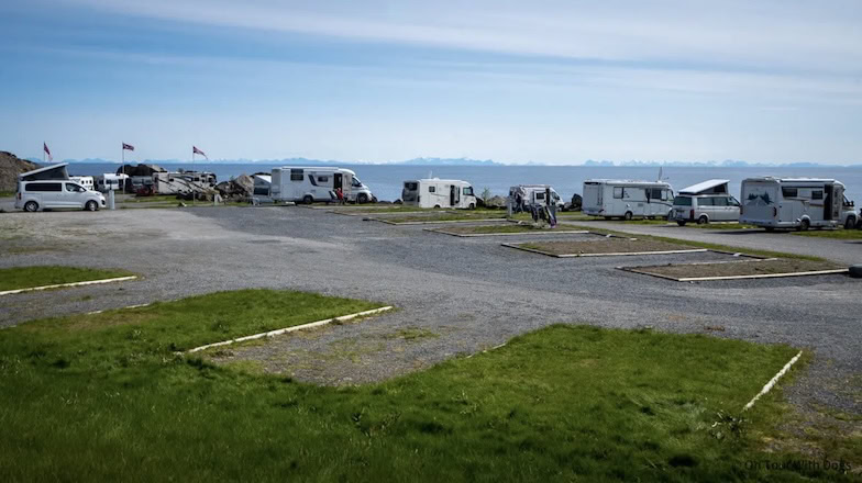 Mehrere Wohnmobile und Wohnwagen stehen auf einem Küstencampingplatz mit Gras- und Schotterparkplätzen. Das Meer und die Berge in der Ferne sind unter einem blauen Himmel zu sehen. Im Hintergrund wehen norwegische Flaggen.
