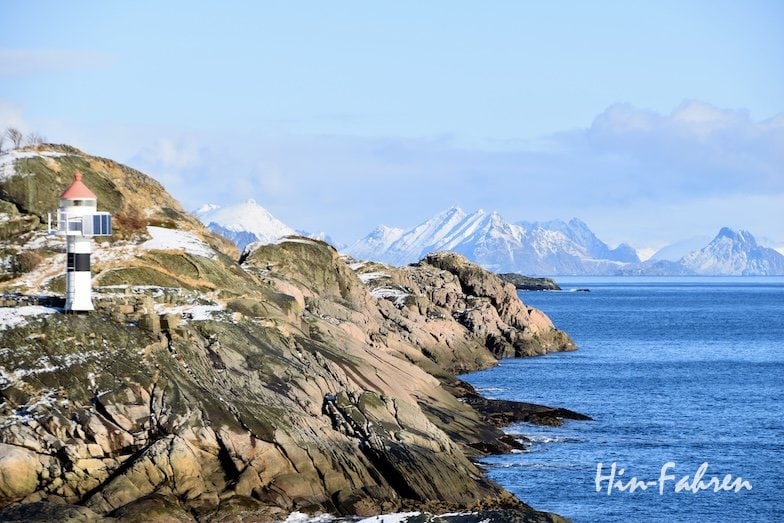 Ein kleiner Leuchtturm steht an einer felsigen, schneebedeckten Küste mit Blick auf ein ruhiges, blaues Meer und ferne, schneebedeckte Berge unter einem teilweise bewölkten Himmel.
