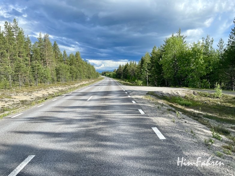 Eine zweispurige Straße erstreckt sich in der Ferne, gesäumt von dichten grünen Wäldern unter einem dramatischen, wolkenverhangenen Himmel mit einigen Sonnenflecken. Die Atmosphäre ist ruhig und heiter.