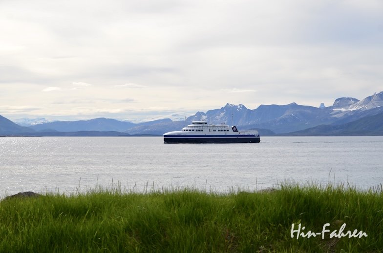 Eine weiß-blaue Fähre fährt über ein ruhiges Gewässer, mit grünem Gras im Vordergrund und fernen Bergen unter einem bewölkten Himmel im Hintergrund. In der unteren rechten Ecke ist der Schriftzug Hin-Fahren zu sehen.