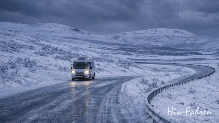 Ein weißes Wohnmobil fährt auf einer kurvenreichen, schneebedeckten Straße durch eine hügelige, winterliche Landschaft in Norwegen unter einem bewölkten Himmel. Die Szene wirkt kalt und abgelegen. In der unteren rechten Ecke stehen die Worte Hin-Fahren.