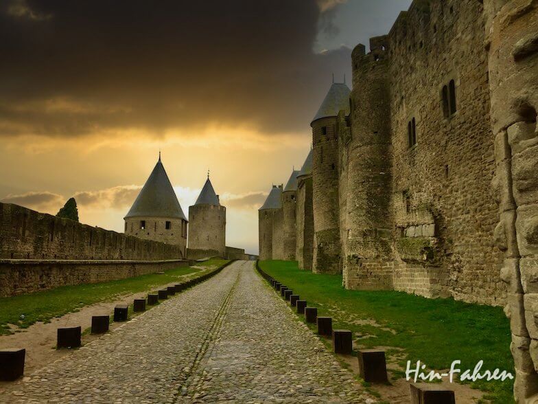 Ein kopfsteingepflasterter Weg führt zwischen hohen Steinmauern und Türmen einer mittelalterlichen Burg bei Sonnenuntergang, mit dramatischen Wolken am Himmel und grünem Gras an den Seiten.