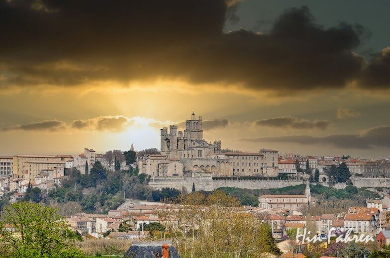 Ein dramatischer Sonnenuntergang mit dunklen Wolken beleuchtet eine historische Kathedrale auf einem Hügel und die umliegenden Gebäude in Béziers, Frankreich, und wirft ein warmes Licht auf die Stadtlandschaft. Die Signatur des Künstlers erscheint in der unteren rechten Ecke.